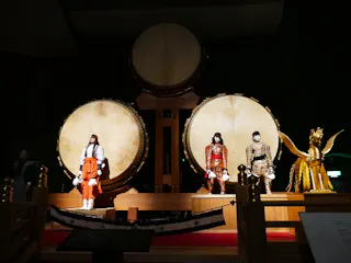 Four traditional Japanese figures in elaborate costumes stand in front of three large taiko drums on a dimly lit stage, with one figure dressed as a golden bird at the far right.
