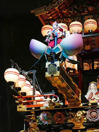 A colorful, ornate Japanese festival float decorated with lanterns, gold accents, and a large lotus blossom. A figure in traditional costume stands atop the float, surrounded by flowers and intricate details.