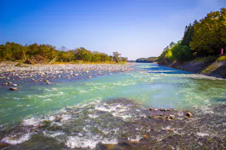 A clear river flows gently between rocky banks and tree-lined shores under a bright blue sky, with sunlight illuminating the greenish-blue water and lush greenery.