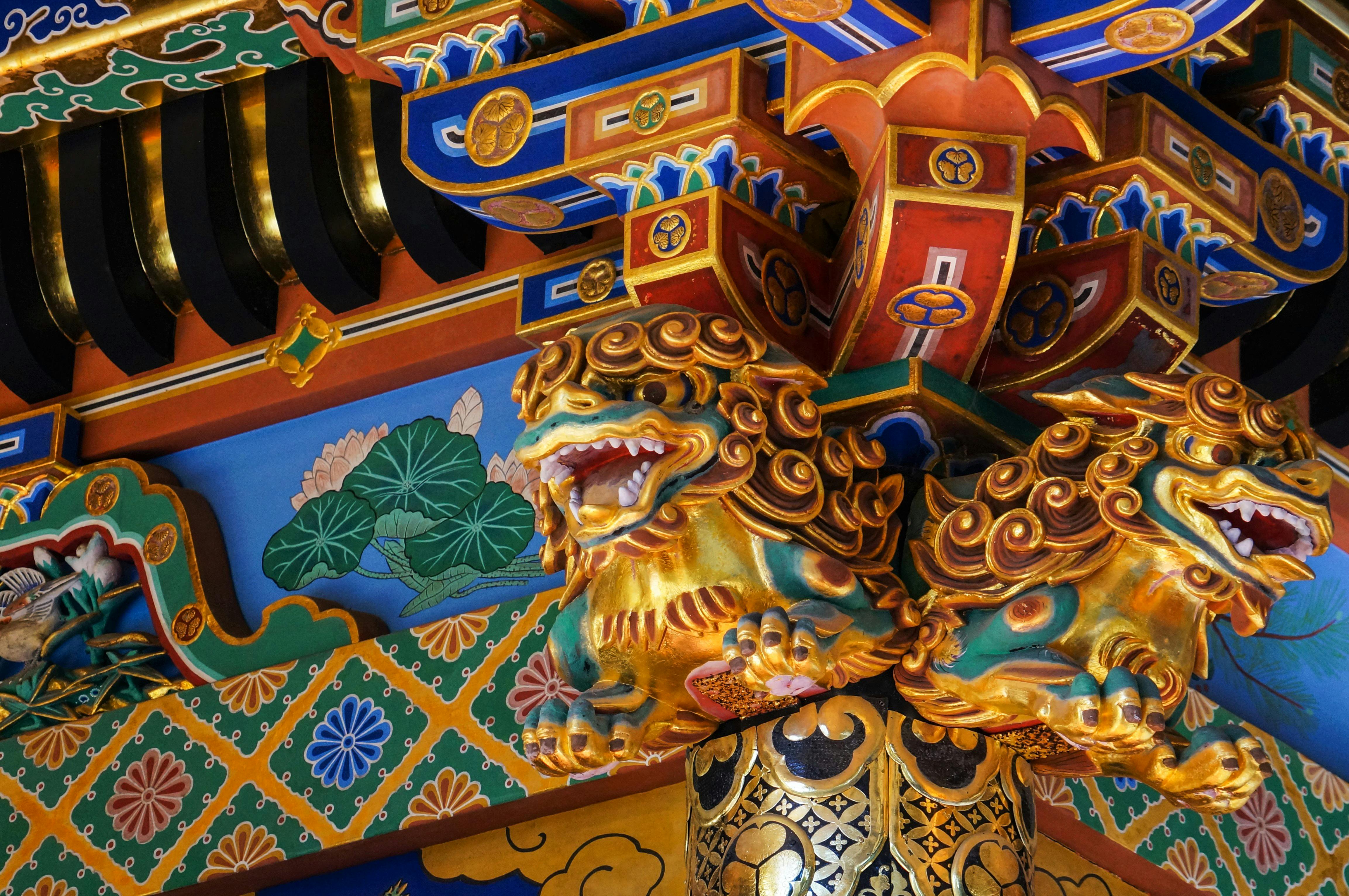 Close-up of an ornate, colorful architectural detail with two golden lion sculptures, intricate patterns, and vibrant floral and geometric designs, typical of traditional East Asian temple or shrine decoration.