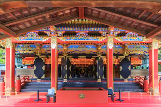 The image shows an ornately decorated Japanese shrine with vibrant red pillars, gold accents, and intricate patterns on the roof. The open structure reveals an inner altar adorned with hanging lanterns and detailed woodwork.