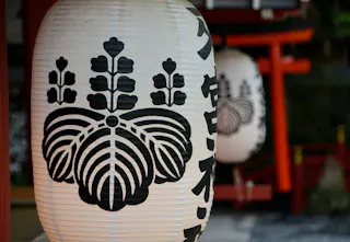 A close-up of a traditional Japanese paper lantern with a black floral design and Japanese characters, hanging outdoors near another lantern and a red torii gate in the background.