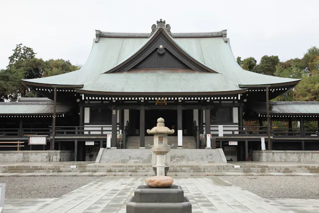A traditional Japanese temple with a large, sloped roof, wooden beams, and a stone lantern in front, surrounded by trees and a paved courtyard.