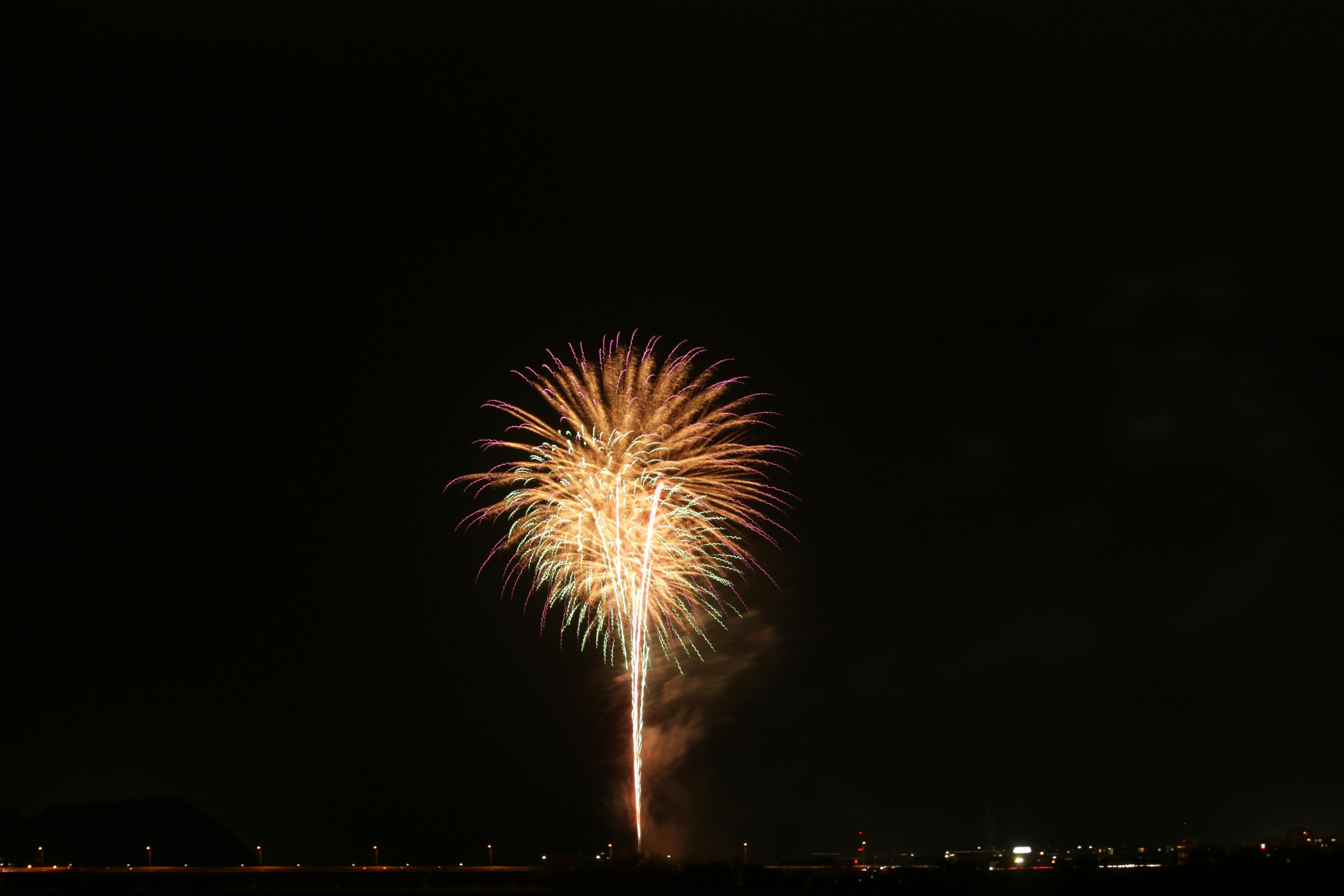 A single bright firework bursts in the night sky, creating yellow and purple streaks above a dark landscape with faint city lights visible along the horizon.