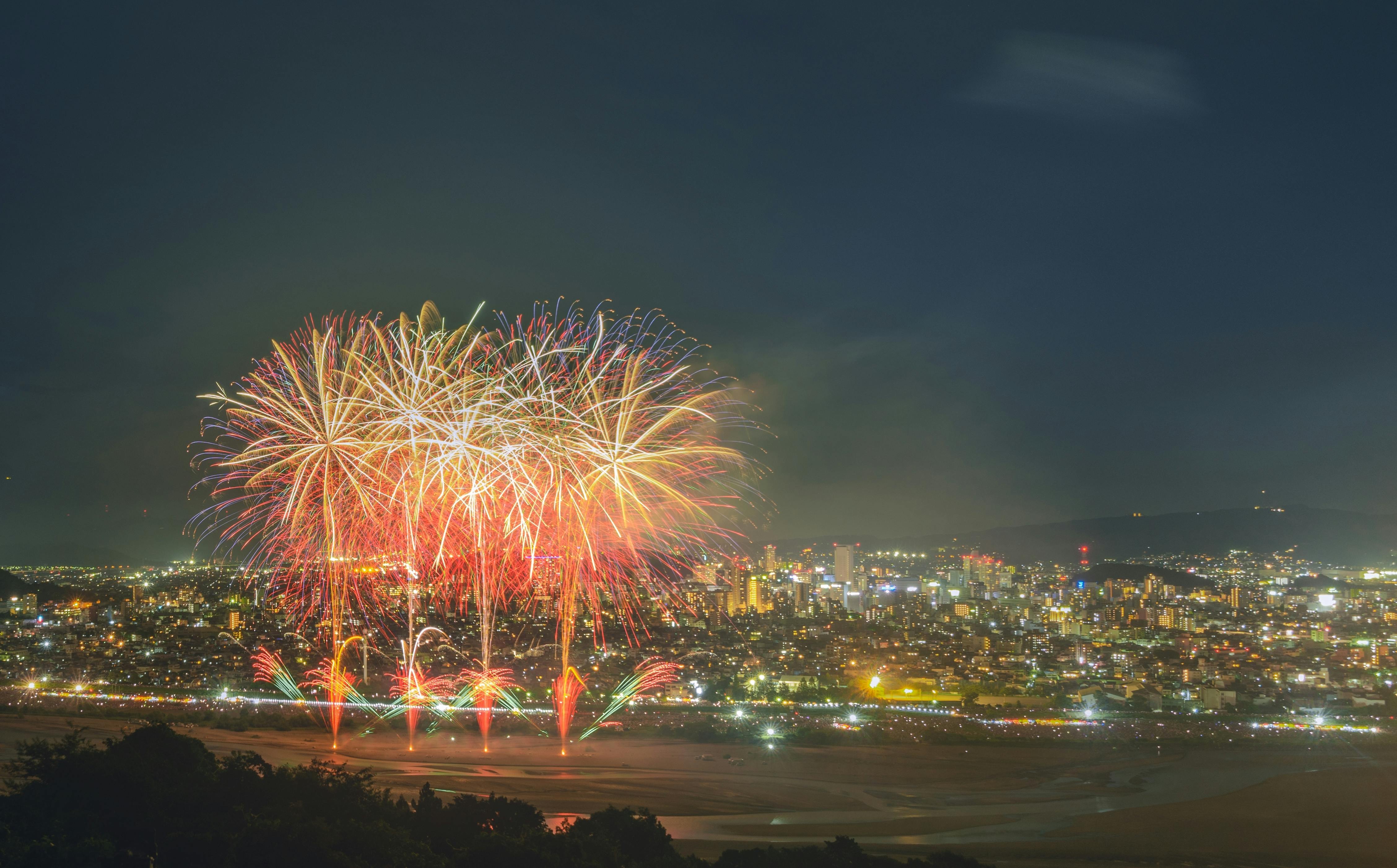 Colorful fireworks burst in the night sky over a brightly lit cityscape, illuminating buildings and streets below, with silhouetted hills in the foreground.