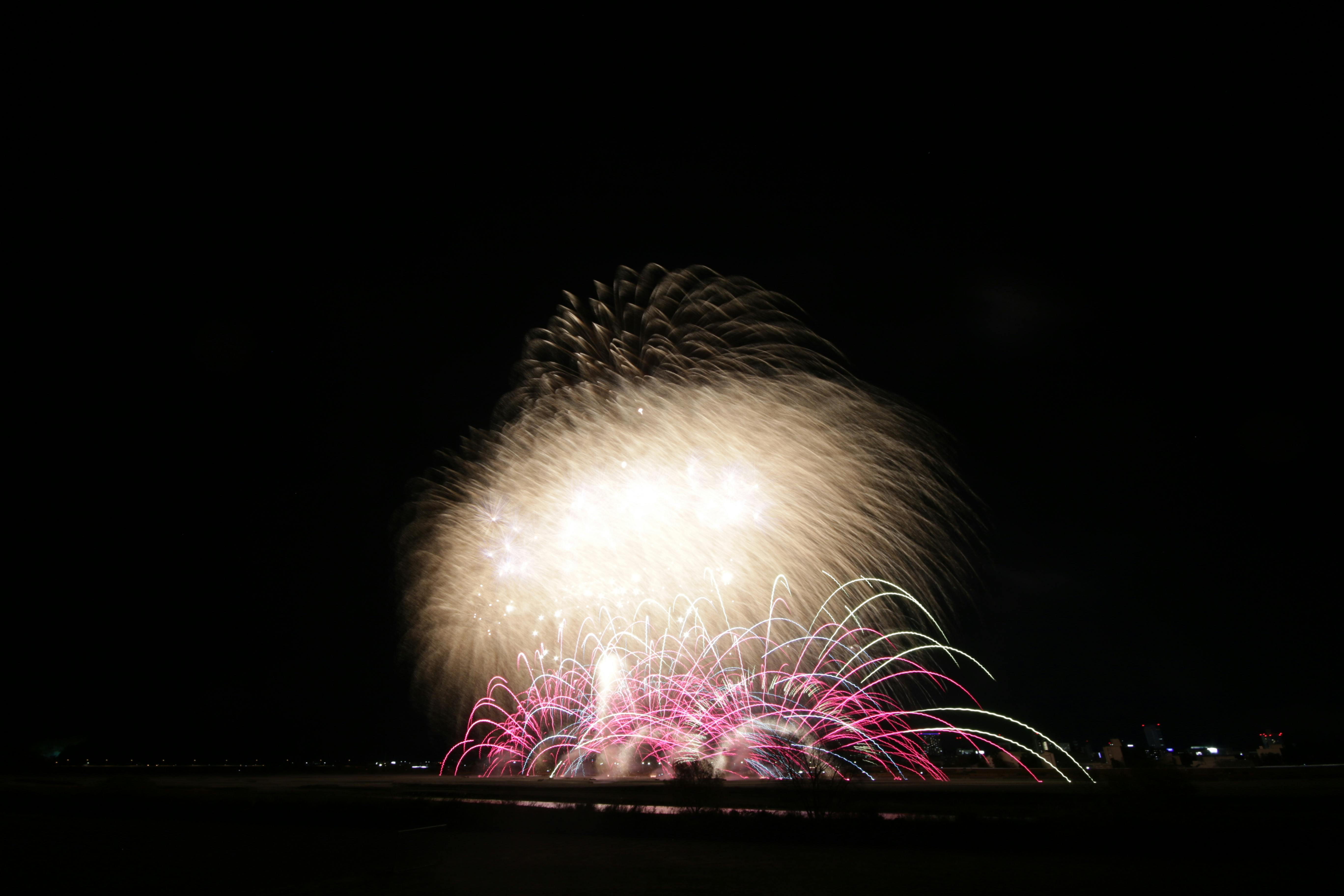 Fireworks light up the night sky, with bright white and red bursts spreading outward in various directions over a dark landscape with faint city lights in the distance.