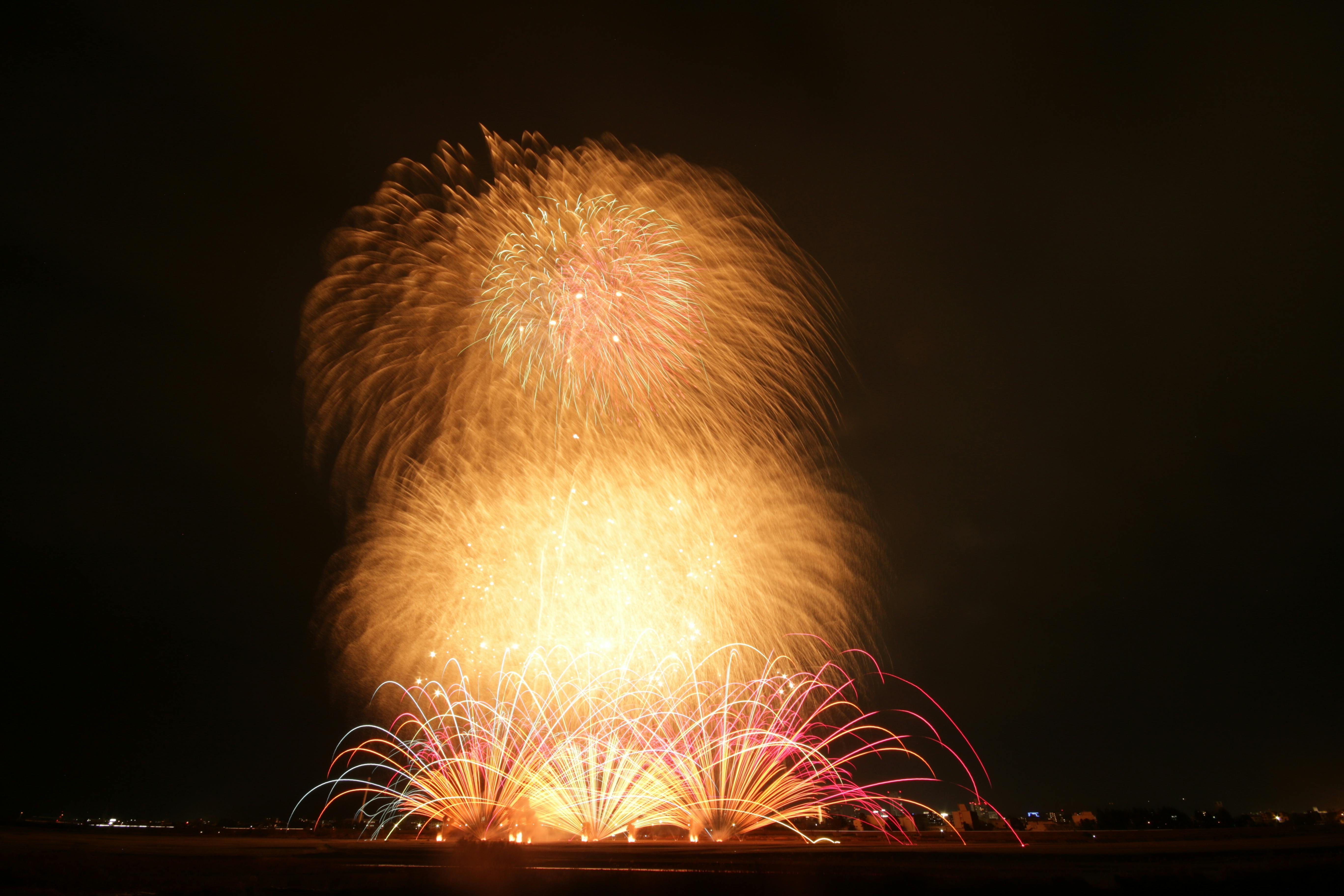A large display of fireworks exploding in the night sky, featuring bright golden and red bursts spreading outward above a dark landscape.
