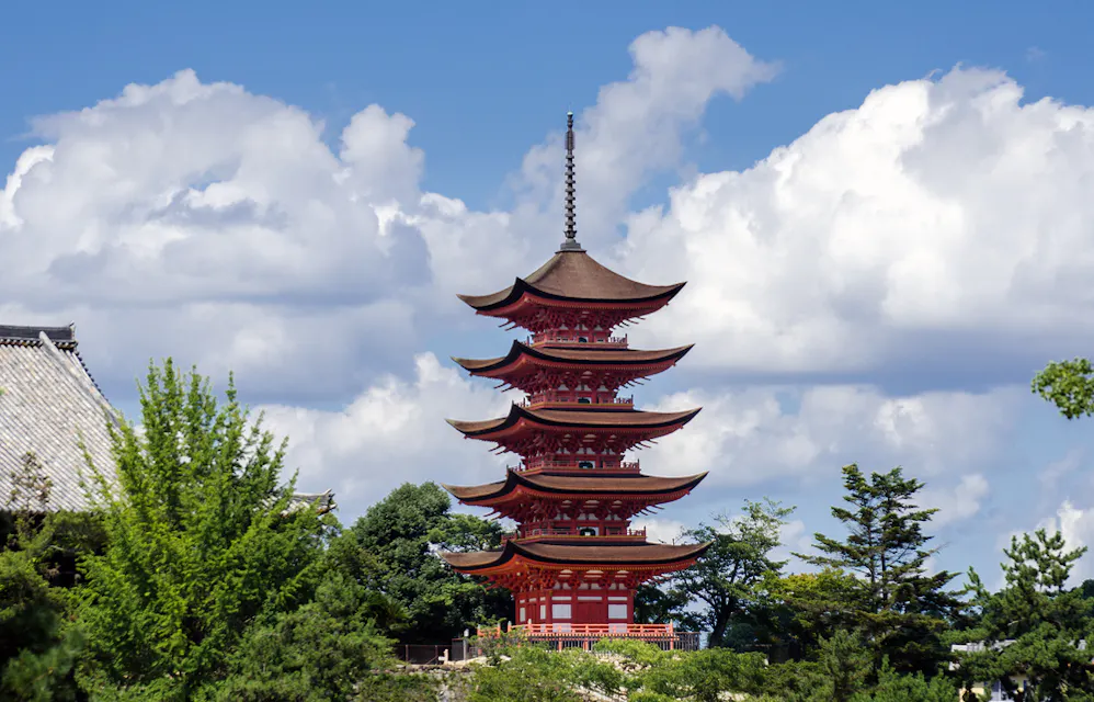 Five-Story Pagoda A five-story red pagoda stands against a backdrop of a bright blue sky with fluffy white clouds. Surrounded by lush green trees, the pagoda features traditional Japanese architectural design with upward-curving eaves on each level.