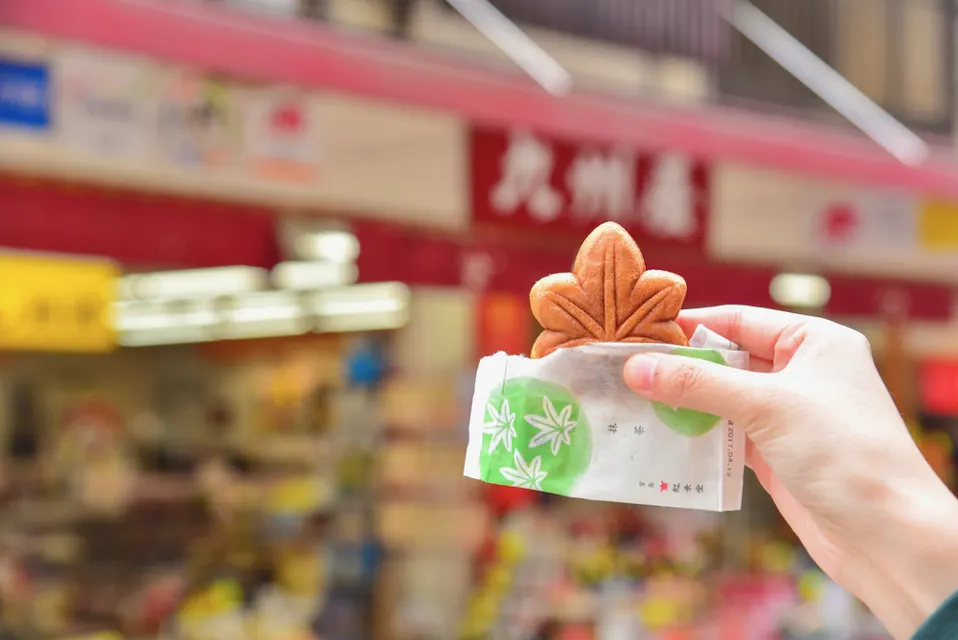 Momiji Manju A hand holds a brown leaf-shaped confectionery wrapped partially in white paper with green leaf patterns. The background features a shop with red and white signage and blurred products, suggesting a bustling retail environment.
