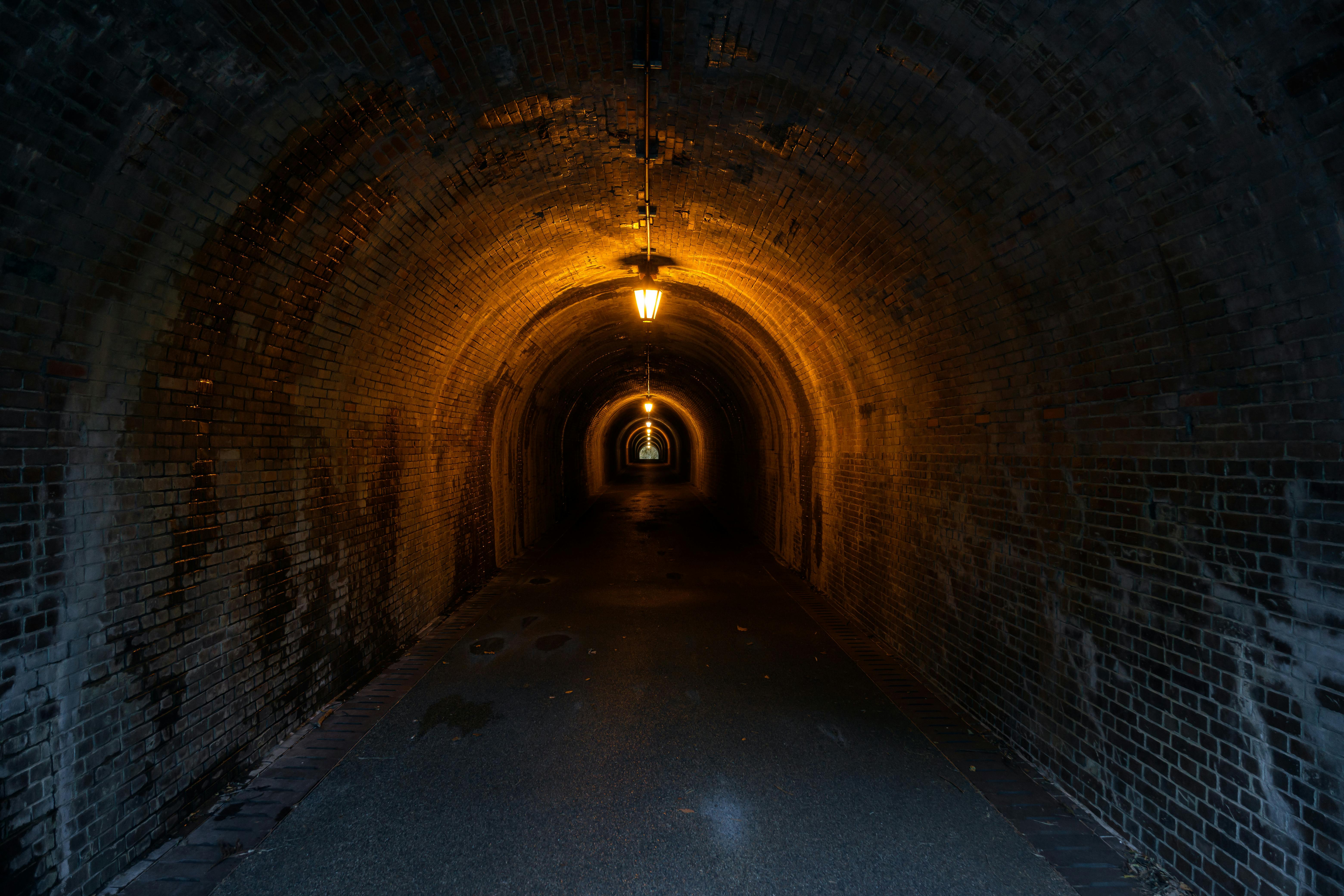 A dimly lit, brick-lined tunnel with glowing yellow lights overhead, creating a sense of depth and leading the viewer’s eye toward the distant, brighter exit at the far end.