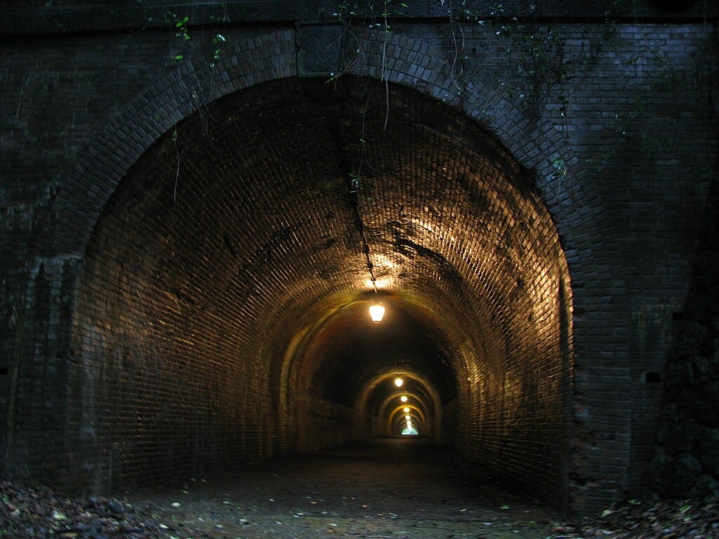 A dimly lit, arched brick tunnel stretches into the distance. Yellow lights line the ceiling, creating a warm glow against the dark, textured walls. Faint natural light is visible at the tunnel’s far end.