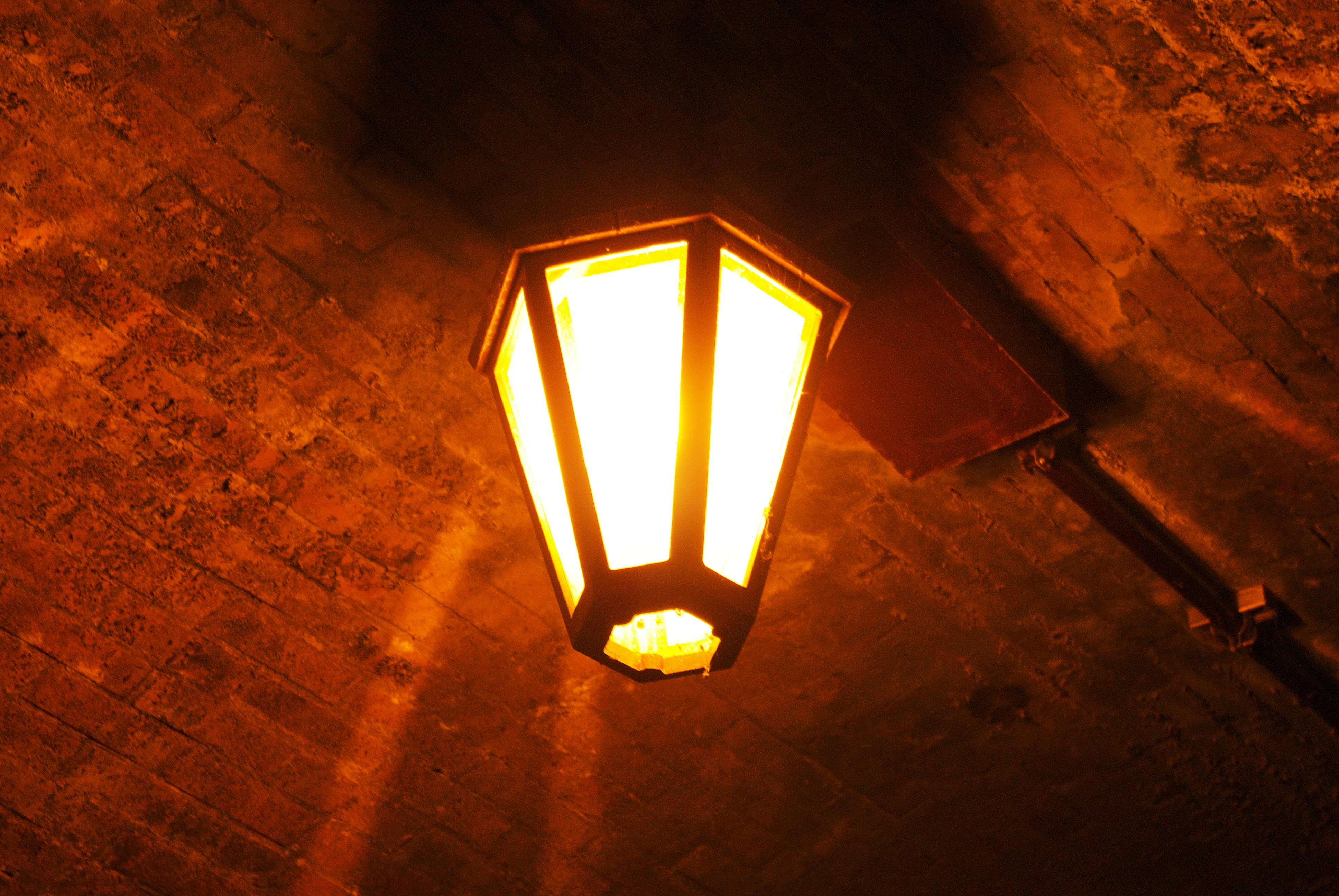 A glowing vintage-style lantern attached to a brick ceiling casts a warm, orange light, creating a cozy and slightly moody atmosphere.