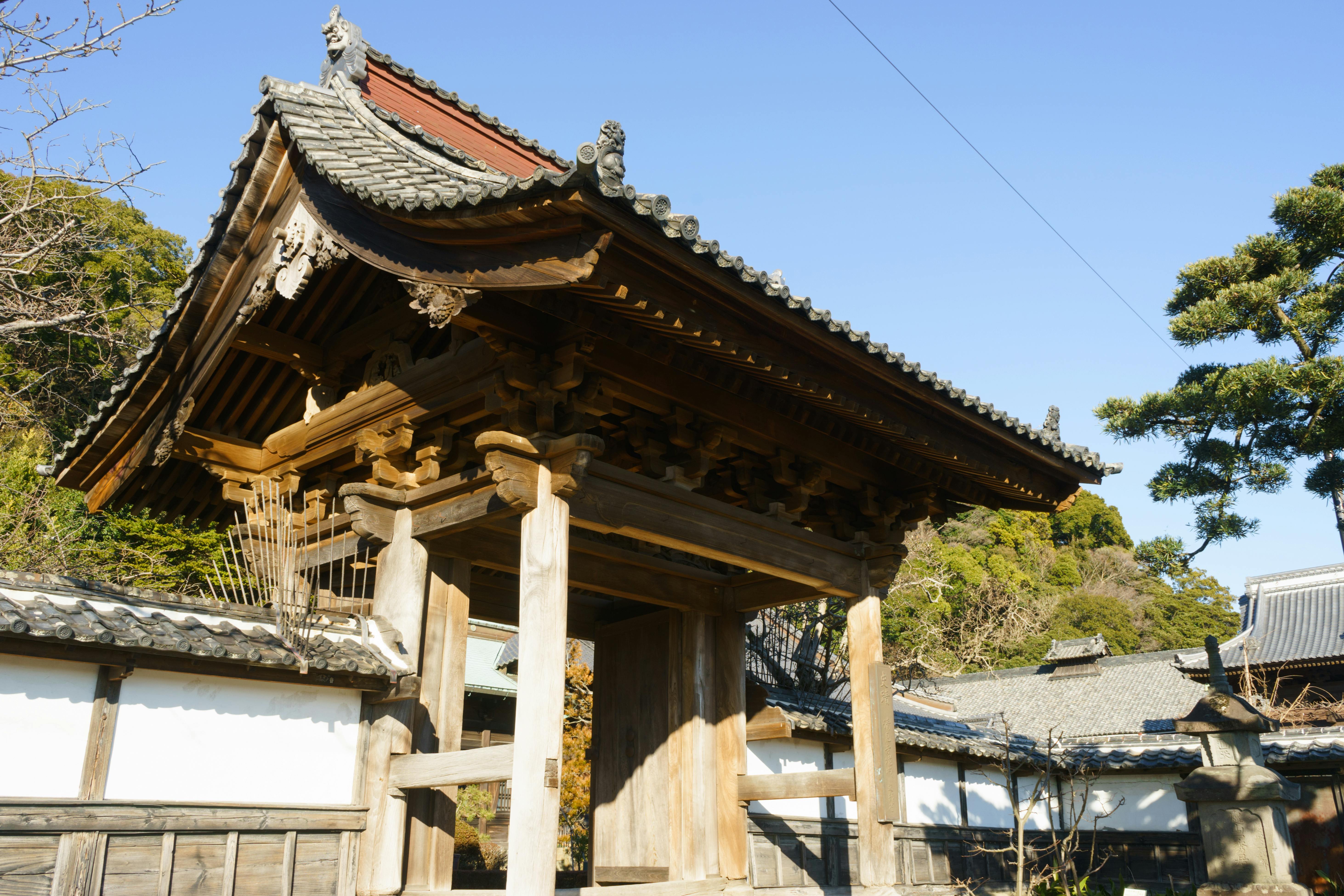 A traditional Japanese wooden gate with ornate roof tiles stands at the entrance of a building complex, surrounded by white walls and trees under a clear blue sky.