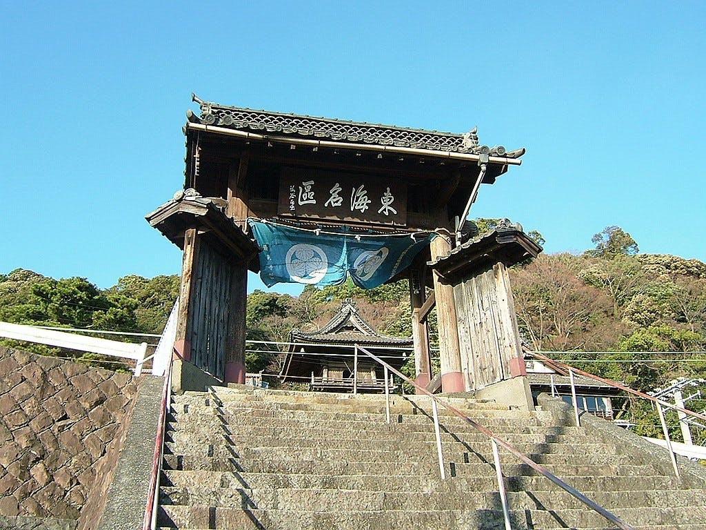 A traditional Japanese temple gate with wooden pillars and a tiled roof stands at the top of a stone staircase, with trees and a blue sky in the background.