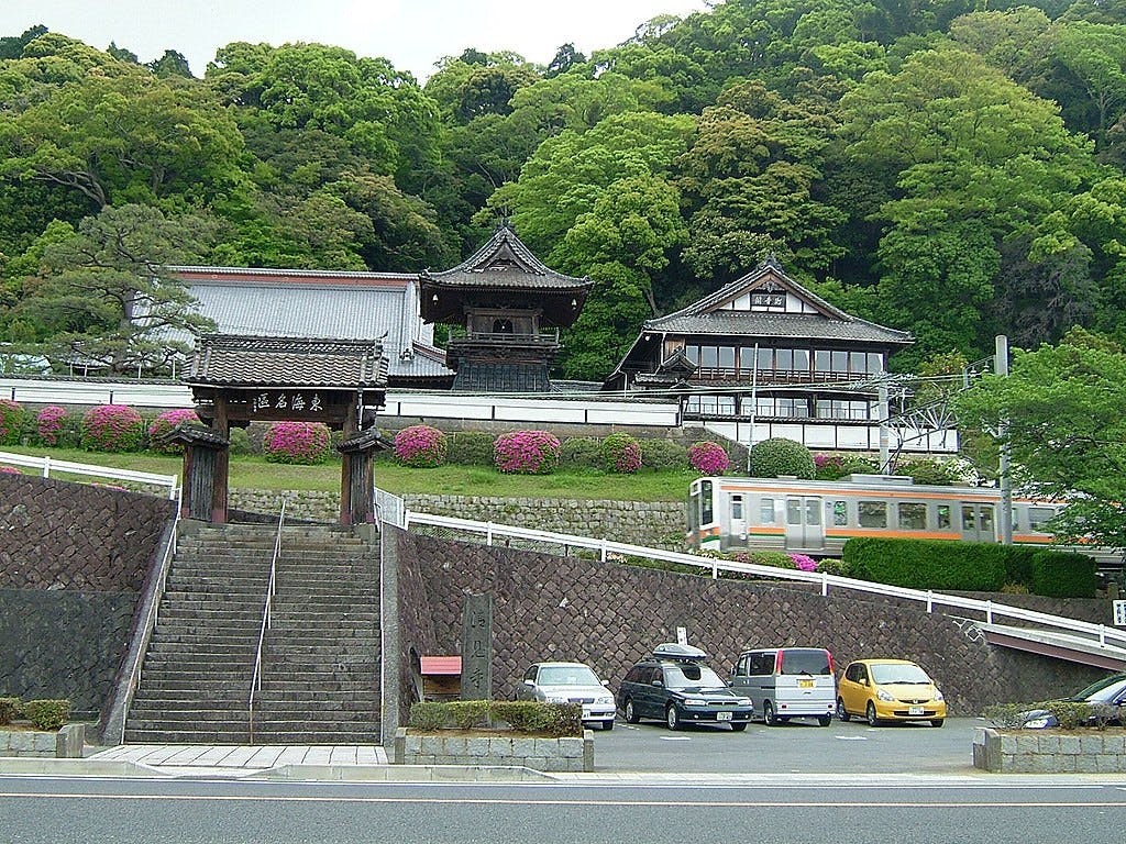 A traditional Japanese temple with sloped roofs sits on a hill, surrounded by green trees and azalea bushes. A small train passes by, and several cars are parked below near stone steps and a wooden gate.