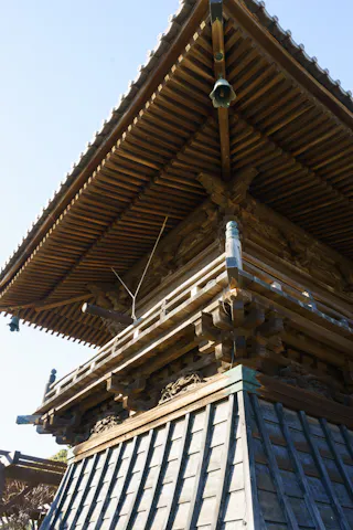 Upward view of the corner of a traditional Japanese wooden temple or pagoda, showing intricate wooden details and a tiled roof under a clear blue sky.