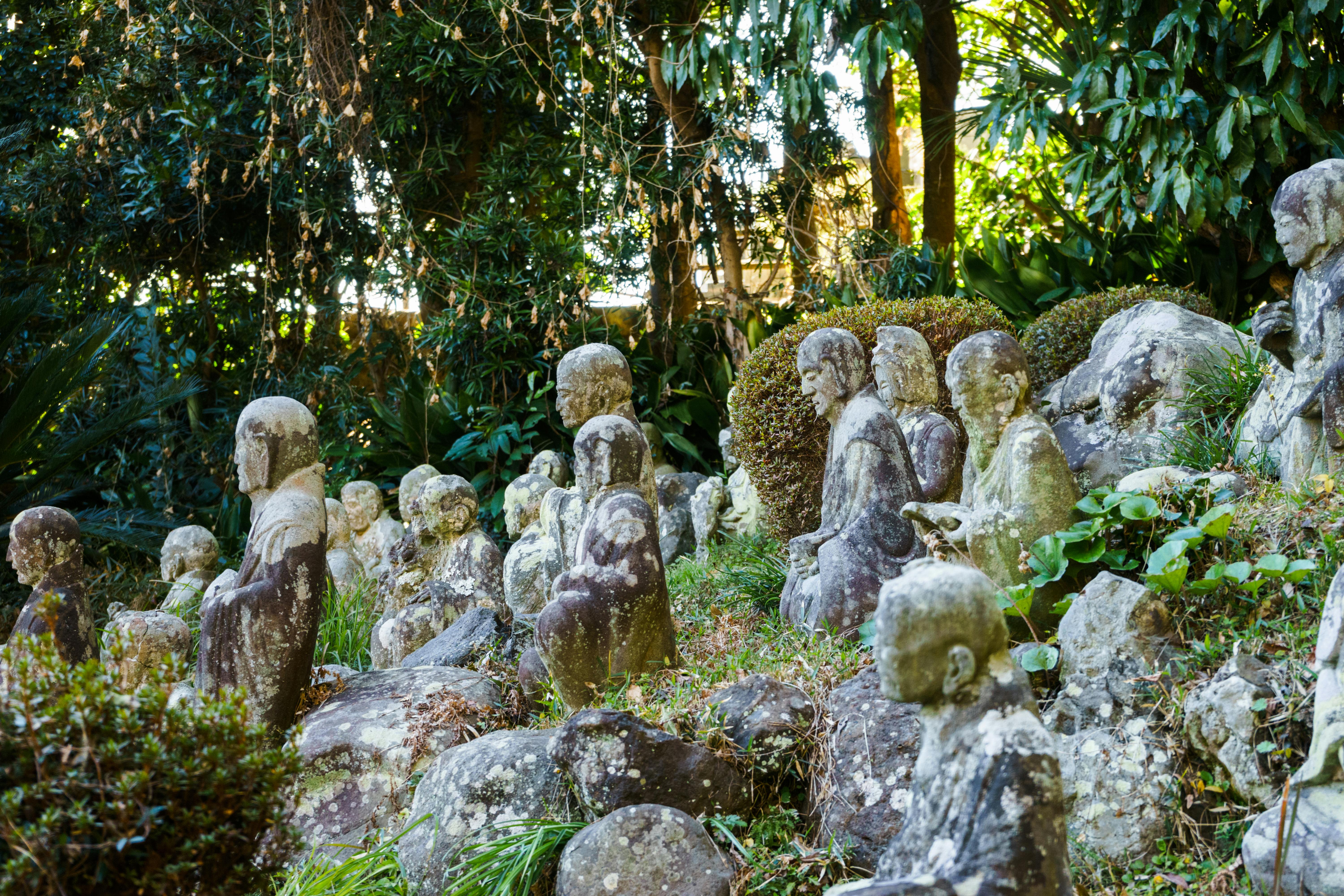 Stone statues of seated figures, weathered by time, are arranged among rocks and greenery in a lush, sunlit garden surrounded by dense trees and plants.