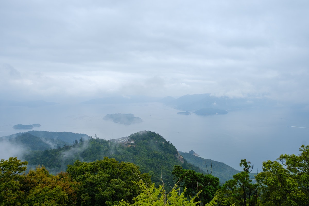 Mount Misen in Miyajima A picturesque view of mist-covered islands and calm waters seen from a lush green hilltop on a cloudy day. The serene landscape features variously sized islands scattered across the tranquil sea, surrounded by gentle fog.