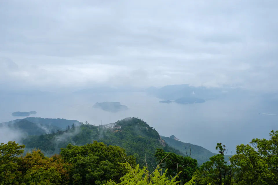 A picturesque view of mist-covered islands and calm waters seen from a lush green hilltop on a cloudy day. The serene landscape features variously sized islands scattered across the tranquil sea, surrounded by gentle fog.