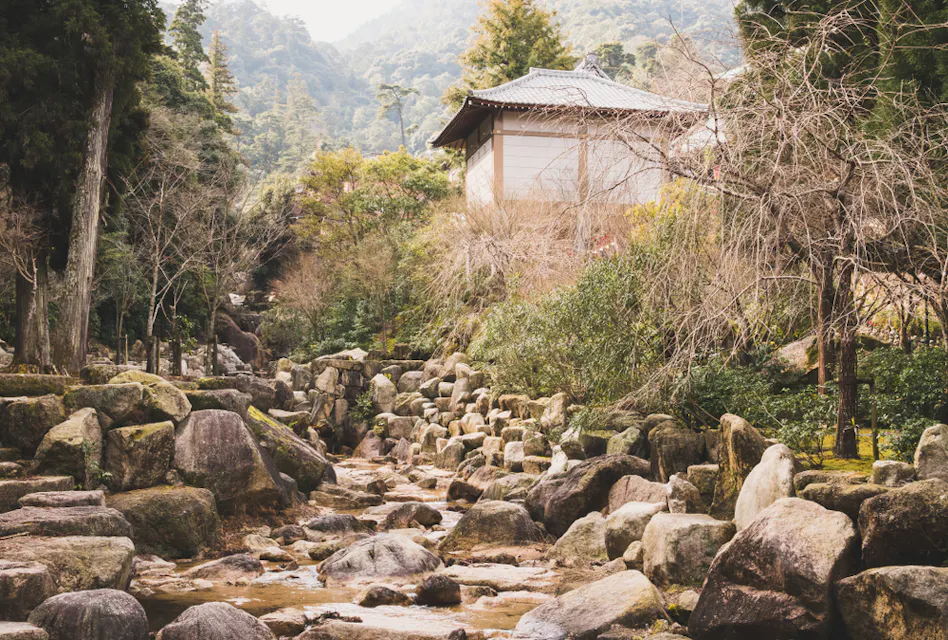 Daisho-in Course A traditional Japanese house sits atop a hill, surrounded by lush greenery and trees. In the foreground, a dry rocky streambed winds through the landscape, leading towards the house. The background features misty, forest-covered mountains.