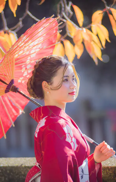 Wearing Kimono A woman in a red kimono holds a red paper umbrella, standing outdoors with autumn leaves in the background. Sunlight softly illuminates her and the colorful foliage.