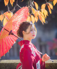 Wearing Kimono A woman in a red kimono holds a red paper umbrella, standing outdoors with autumn leaves in the background. Sunlight softly illuminates her and the colorful foliage.