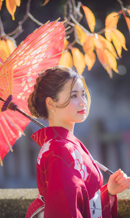 Wearing Kimono A woman in a red kimono holds a red paper umbrella, standing outdoors with autumn leaves in the background. Sunlight softly illuminates her and the colorful foliage.