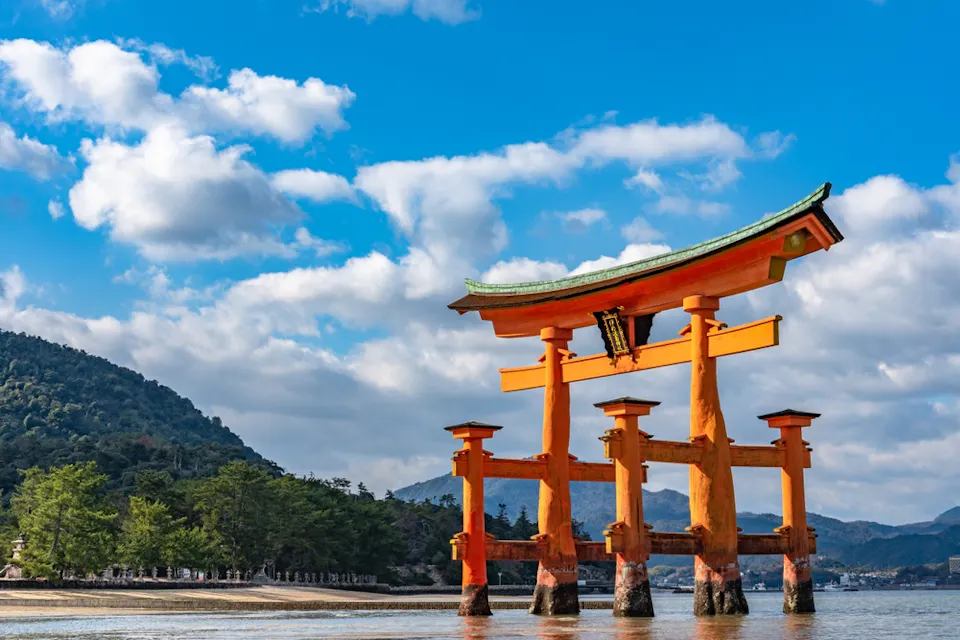 Floating Torii Gate A large, iconic torii gate painted in bright orange stands in shallow waters with mountains and forested coastline in the background. The sky is filled with scattered clouds on a sunny day, casting reflections on the water's surface.