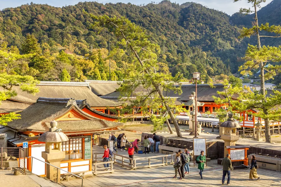 Itsukushima Shrine A group of people visit a traditional Japanese shrine with red and beige buildings. The shrine is set against a backdrop of lush green mountains under a clear sky. Tall pine trees are scattered around the area, enhancing the serene atmosphere.