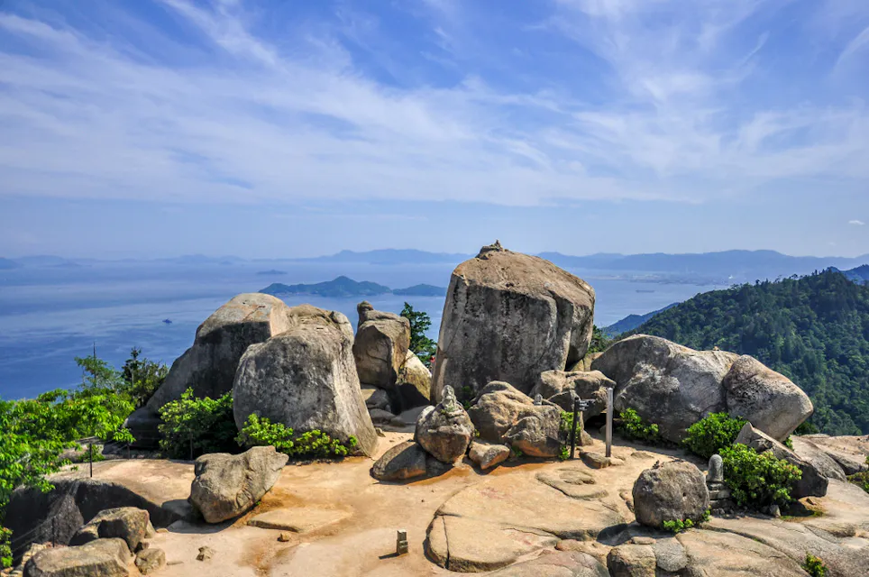 Mount Misen A scenic view from a mountaintop featuring large boulders in the foreground, surrounded by lush greenery. Beyond the rocks, there's an expansive view of the calm blue sea and distant islands under a clear sky. The horizon blends seamlessly with the sky.