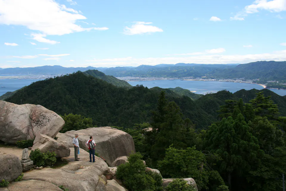 Mount Misen Two people stand atop a rocky outcrop, overlooking a vast landscape of wooded hills and distant water. Below them, dense greenery extends towards the horizon, meeting the blue sky dotted with scattered clouds. The view is expansive and serene.