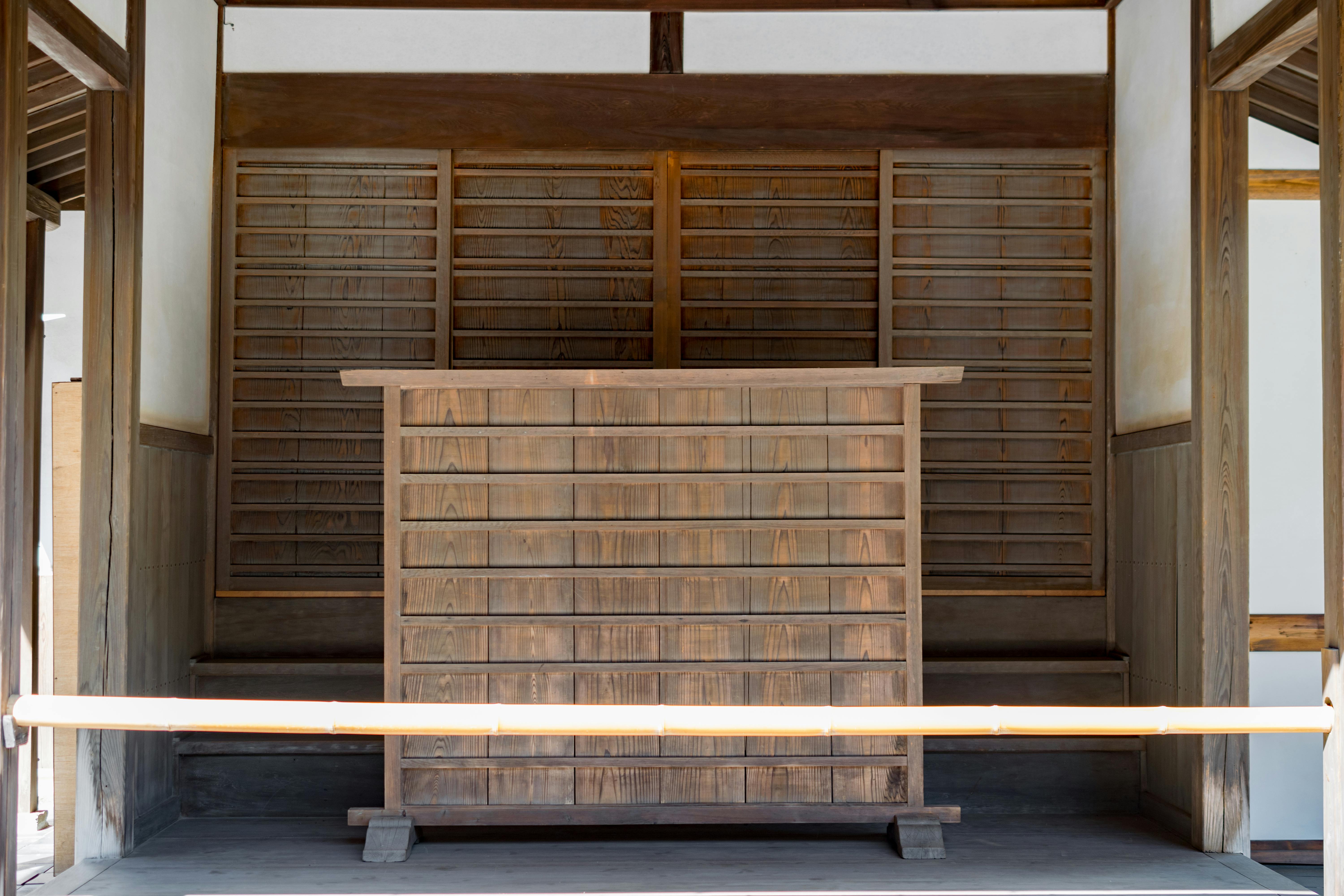 A traditional Japanese wooden reception counter stands in front of sliding wooden doors in a minimalist, sunlit room with wooden walls and a bamboo railing.