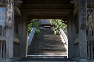 A view through a wooden temple gate reveals a wide stone staircase leading up to a traditional Japanese building, surrounded by greenery in the background.