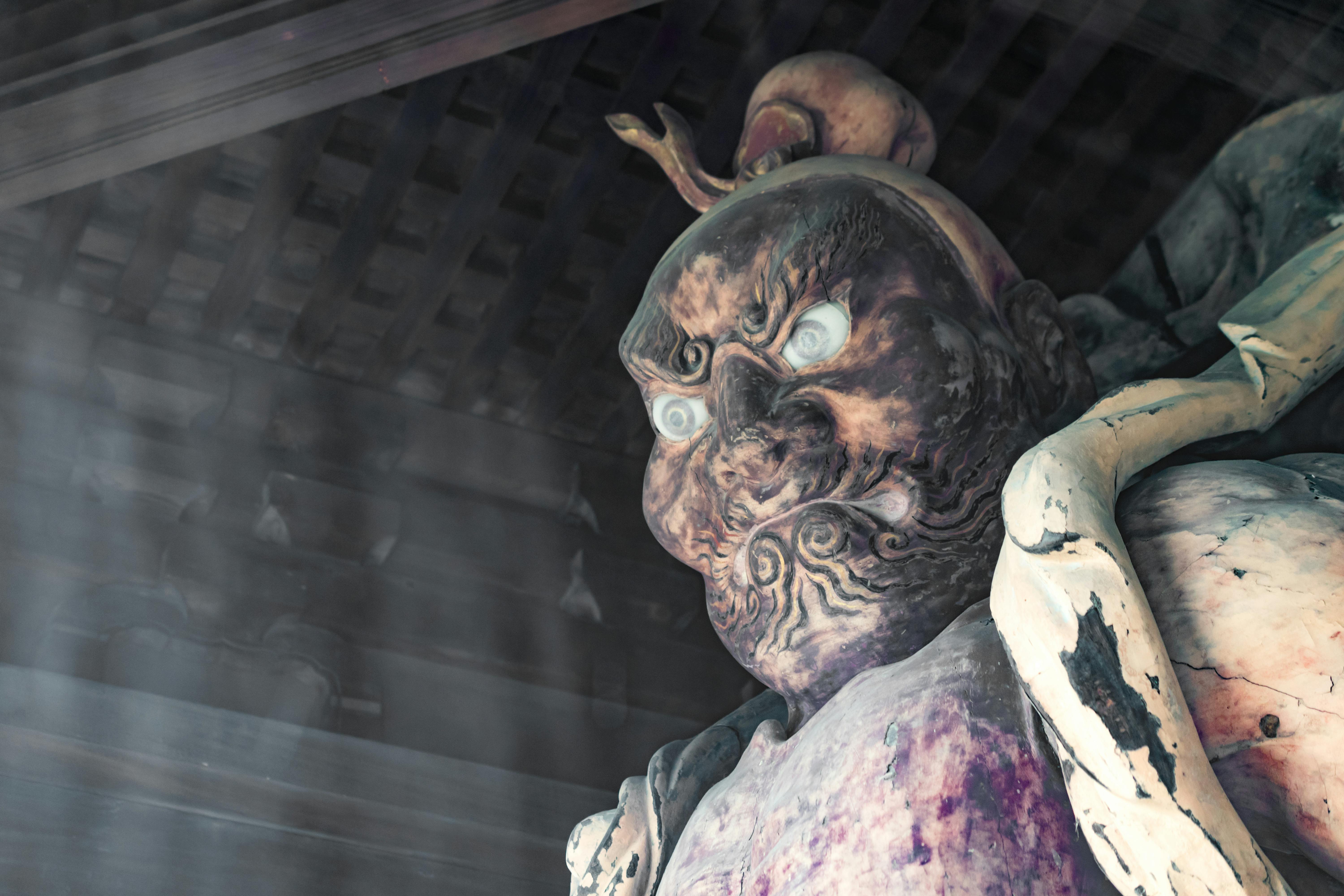 A close-up of a fierce, weathered statue with bulging eyes and an intense expression, partially illuminated by soft light inside a dark wooden structure. The statue has intricate facial details and is adorned with large, flowing ornaments.