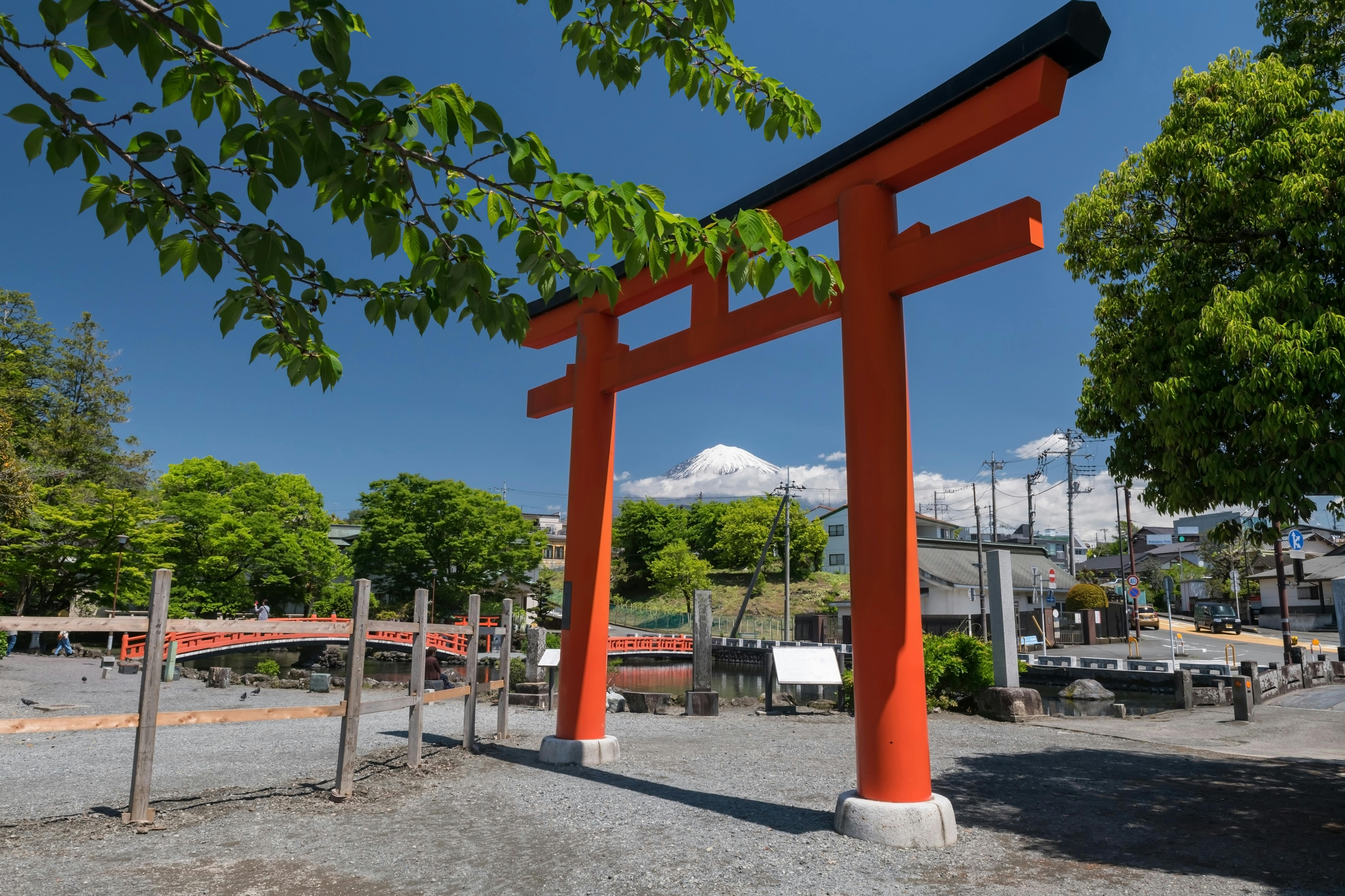 A large red torii gate stands in the foreground, with green trees and a red bridge nearby. Mount Fuji, partly covered in snow, is visible in the background under a clear blue sky.