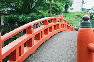 A close-up view of a traditional red arched bridge with railings, surrounded by lush green trees and grass on a cloudy day.