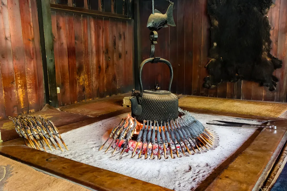 Mountain Lodges or Huts A traditional Japanese irori (sunken hearth) with a kettle hanging over an open fire is surrounded by skewered fish being grilled. The hearth is set in a wooden-floored room with a reddish-brown wooden wall and an animal pelt mounted on the wall.