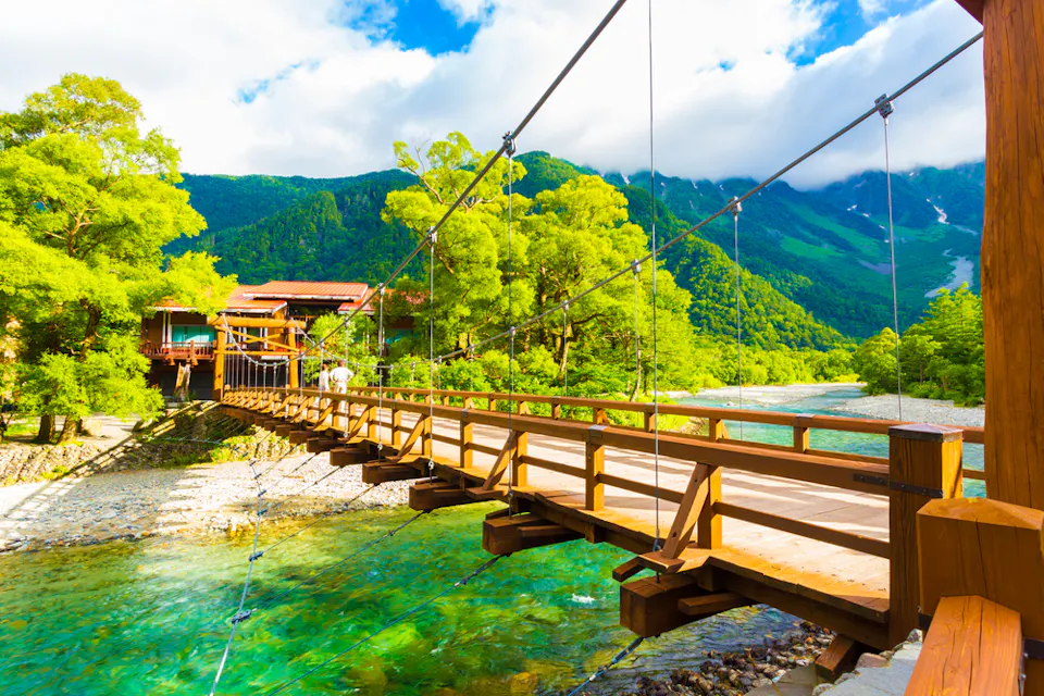 Kappa Bridge A wooden suspension bridge stretches over a clear, turquoise river, surrounded by lush green trees and vibrant mountains. Bright sunlight highlights the fresh greenery and the traditional buildings near the bridge. The sky is partly cloudy.