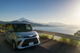 A silver car is parked by a guardrail overlooking the ocean, with mountains and Mount Fuji visible in the background under a clear blue sky.
