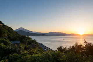A scenic view of the ocean at sunset with lush green hills in the foreground, a road along the shoreline, and Mount Fuji visible in the distance under a clear sky.