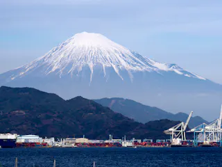 Snow-capped Mount Fuji rises in the background, with a busy port and cranes along the waterfront in the foreground under a clear sky.