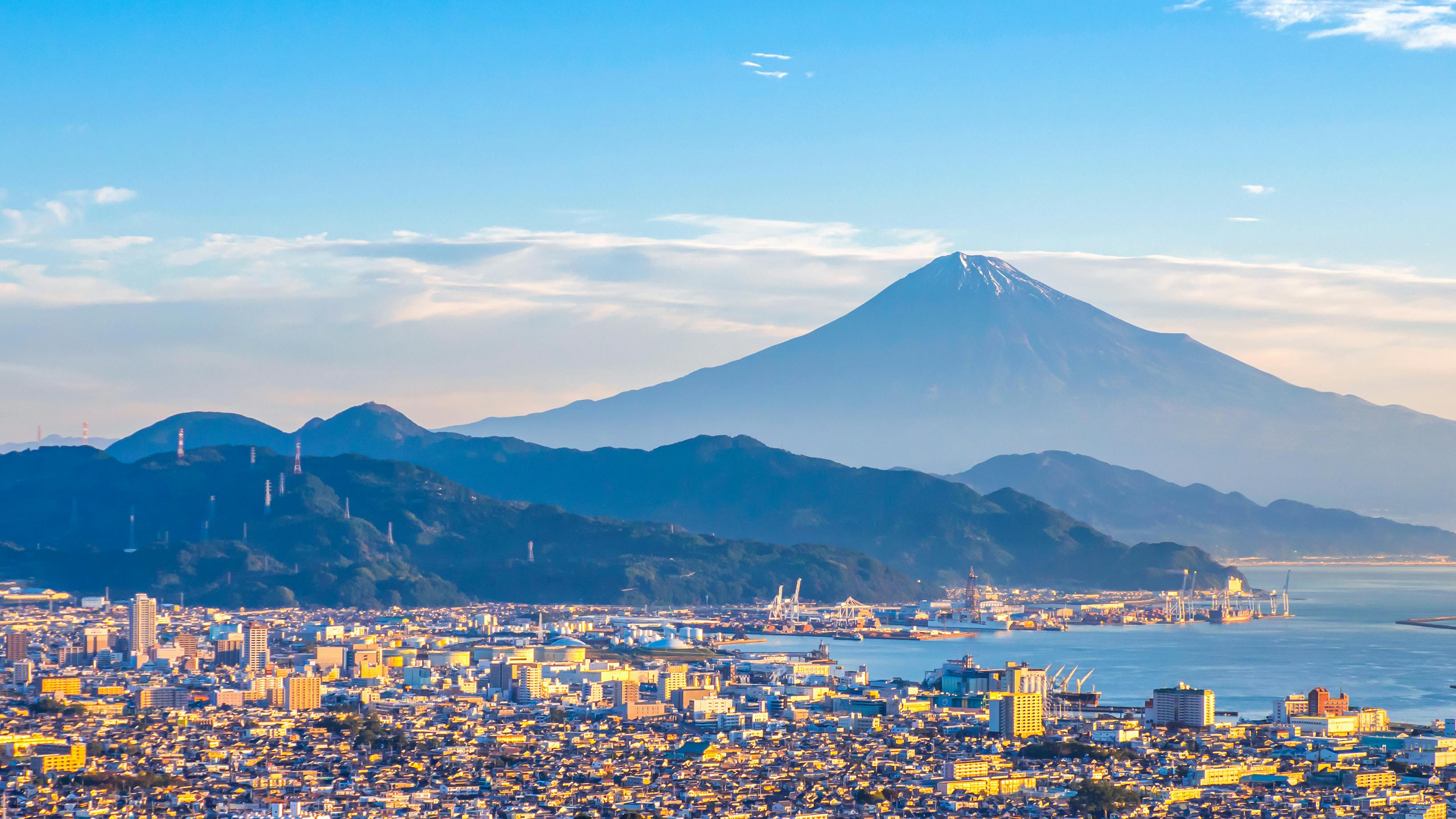 A vibrant cityscape sits beneath a clear sky, with Mount Fuji’s snow-capped peak and green foothills rising majestically in the background.