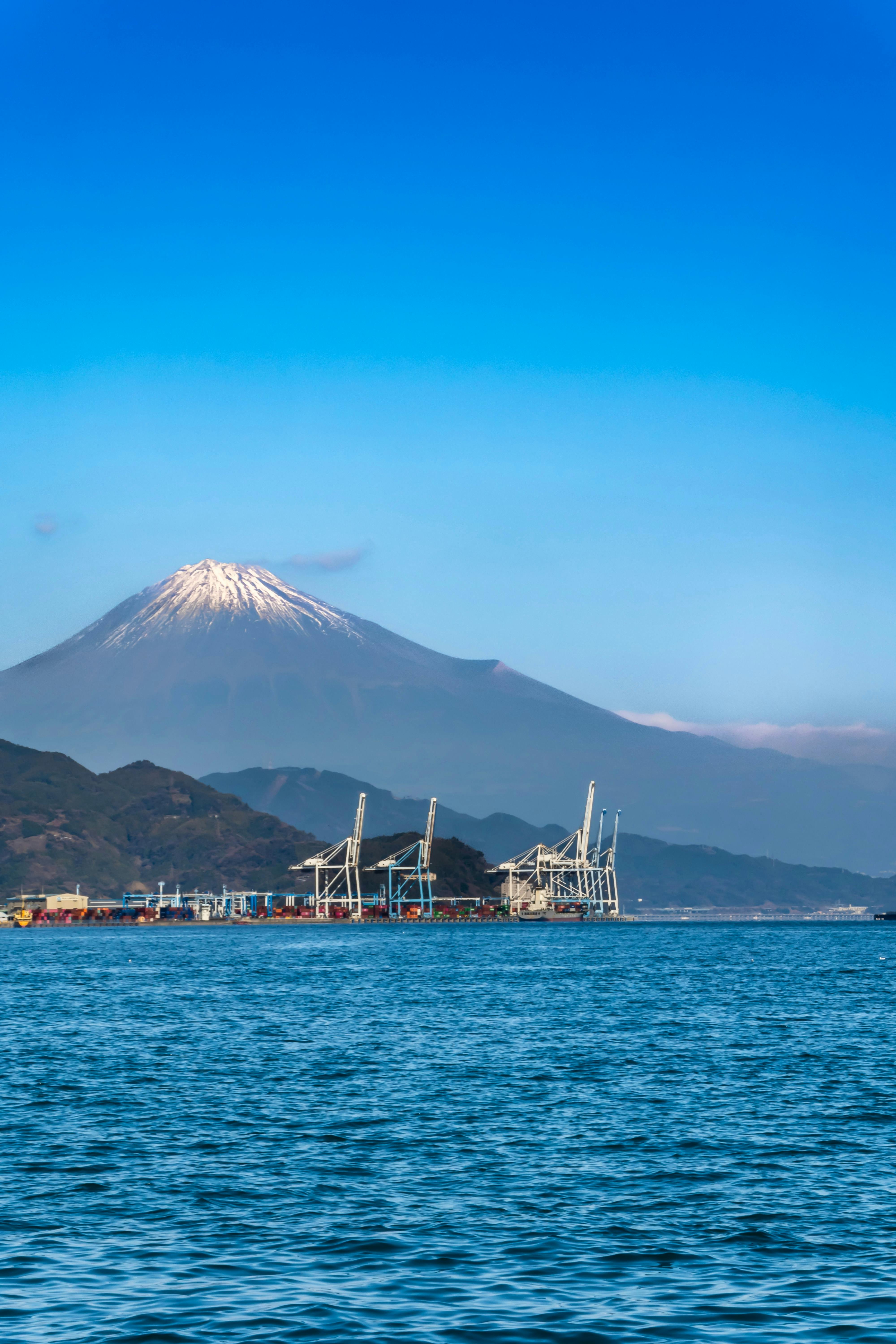 Mount Fuji with a snow-capped peak rises in the background above a port with cranes and buildings along the waterfront, under a clear blue sky and calm water in the foreground.