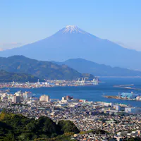 Shimizu Port A clear view of Mount Fuji with a snow-capped peak, towering above a coastal city with ports, buildings, and green hills under a blue sky.