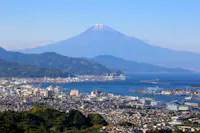 A clear view of Mount Fuji with a snow-capped peak, towering above a coastal city with ports, buildings, and green hills under a blue sky.