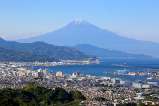 A clear view of Mount Fuji with a snow-capped peak, towering above a coastal city with ports, buildings, and green hills under a blue sky.
