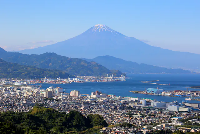 A clear view of Mount Fuji with a snow-capped peak, towering above a coastal city with ports, buildings, and green hills under a blue sky.