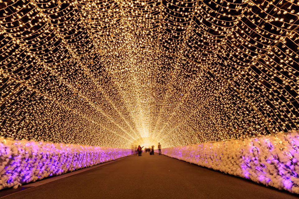 A tunnel illuminated with golden lights, creating an arching, starry ceiling effect. The pathway is adorned with white and purple flower arrangements on both sides. People are seen in the distance, silhouetted by the bright lights, walking through the tunnel.