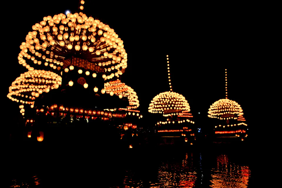 Illuminated structures with numerous glowing lights float on a dark body of water at night. The reflections of the lights shimmer on the water, creating a mesmerizing and festive atmosphere. The scene is bright with a sense of celebration and festivity.
