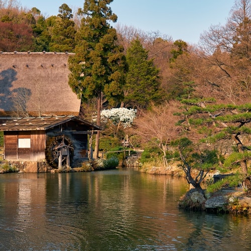 Higashiyama Zoo and Botanical Garden A serene Japanese garden features a traditional wooden watermill by a calm pond. The scene is framed by lush green trees and a thatched-roof building in the background. The soft sunlight enhances the tranquil atmosphere.